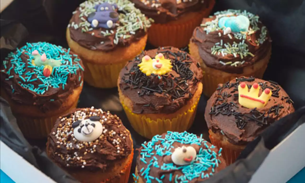 Syv muffins med gyllenbrun bunn, mørk sjokoladeglasur, fargerikt strøssel og små figurer lagt i en hvit eske med sort papir.