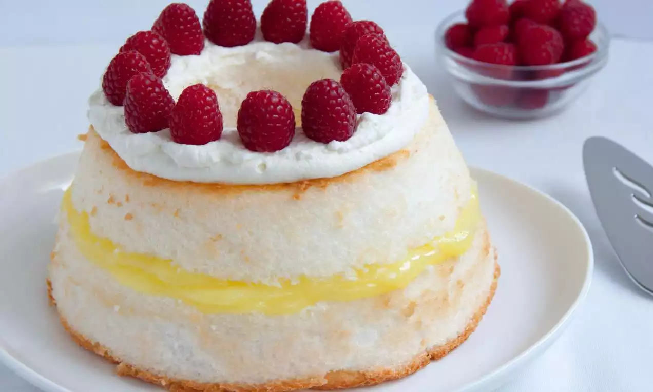 Gâteau aérien aux bords dorés et garniture citronnée, couronné de crème blanche et de framboises rouges sur une assiette blanche