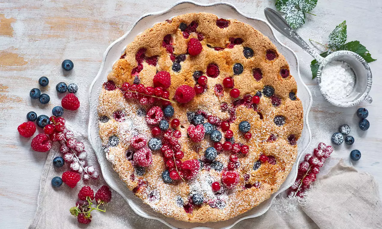 Koláč s chrumkavou zlatohnedou kôrkou, posiaty červenými a modrými bobuľami, ľahko posypaný práškovým cukrom na ozdobnom bielom tanieri