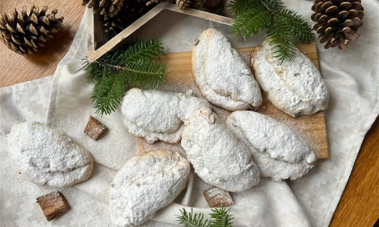 Mini štoly oválneho tvaru, jemne zlatisté, bohato posypané bielym práškovým cukrom na drevenej doske s ihličím a šiškami