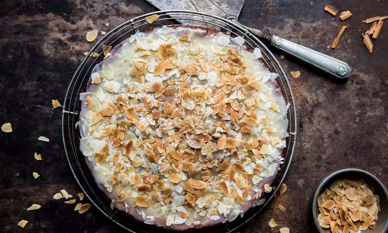 Morotskaka con glassa lucida bianca, coperta di scaglie di mandorle dorate e briciole su una griglia circolare sopra un tavolo scuro