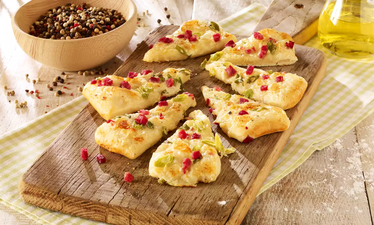 Triangoli dorati di pasta morbida guarniti con pezzetti di prosciutto e erbe verdi su un tagliere di legno accanto a pepe e olio