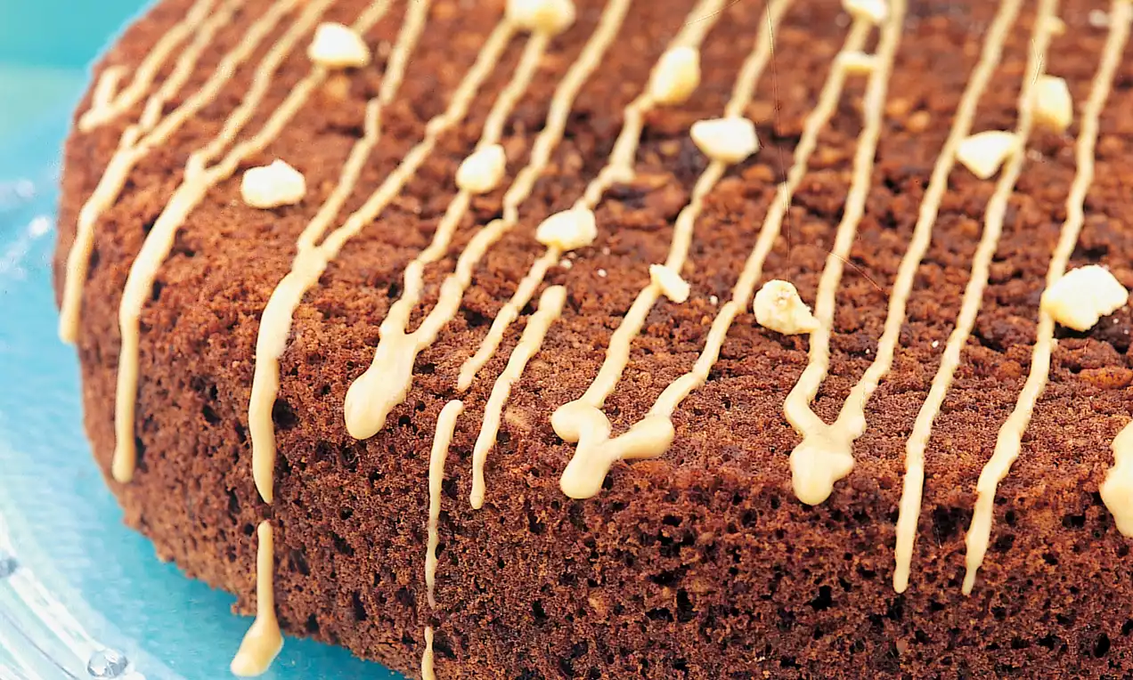 Yoğun kahverengi dokulu kek, üzerine beyaz çikolata şeritleri ve fındık parçaları serpilmiş, cam bir tabakta sunulmuş