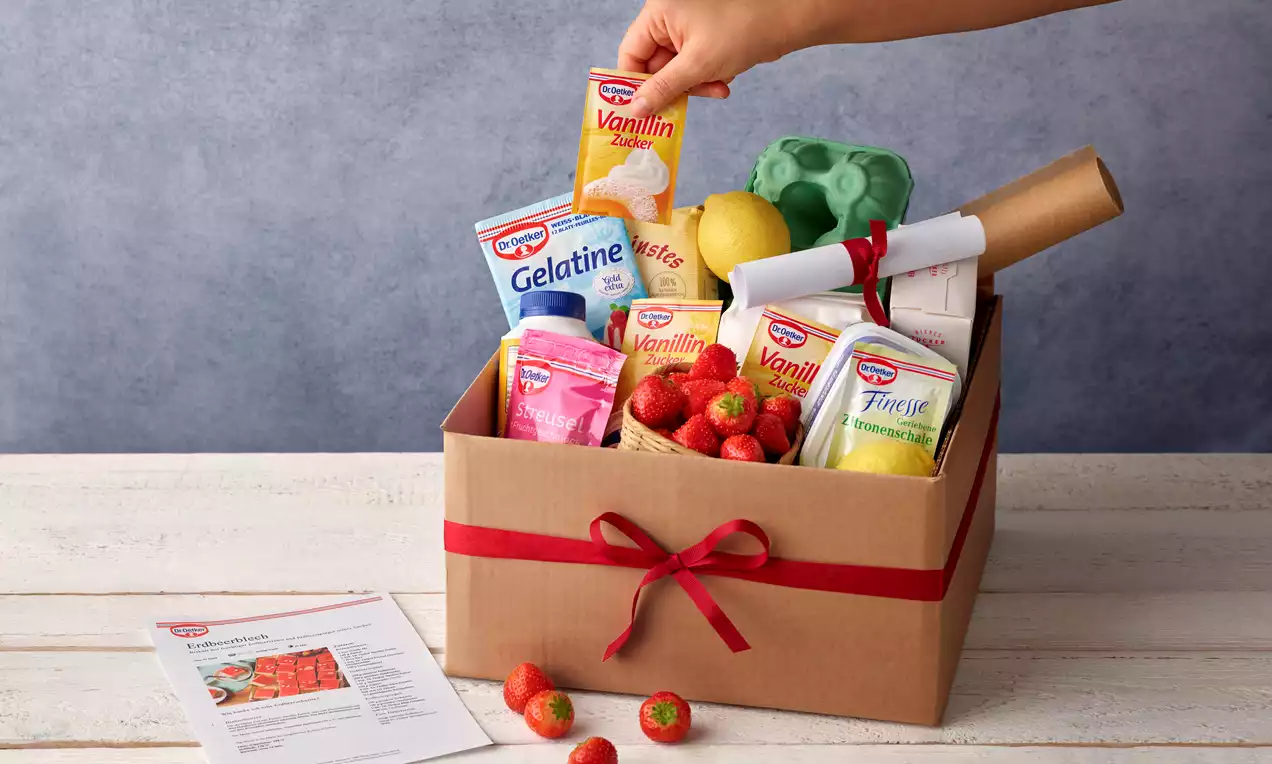 Eine braune Box mit roter Schleife, gefüllt mit Backzutaten wie Vanillezucker, Gelatine, Erdbeeren, Zitrone und Rezeptrolle, daneben frische Erdbeeren