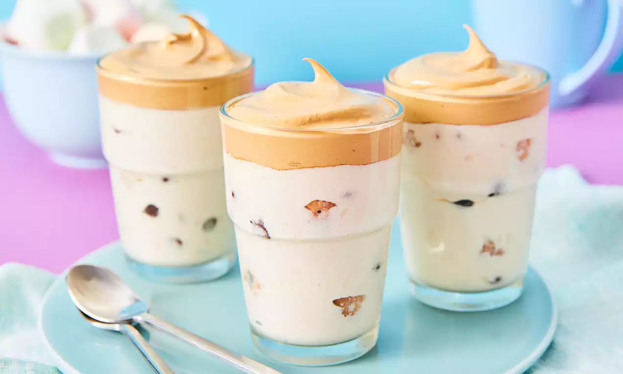 Schichtdessert aus cremiger weißer Füllung mit Schokoladenstücken und goldbrauner Dalgona-Coffee-Schicht in Gläsern auf blauem Teller