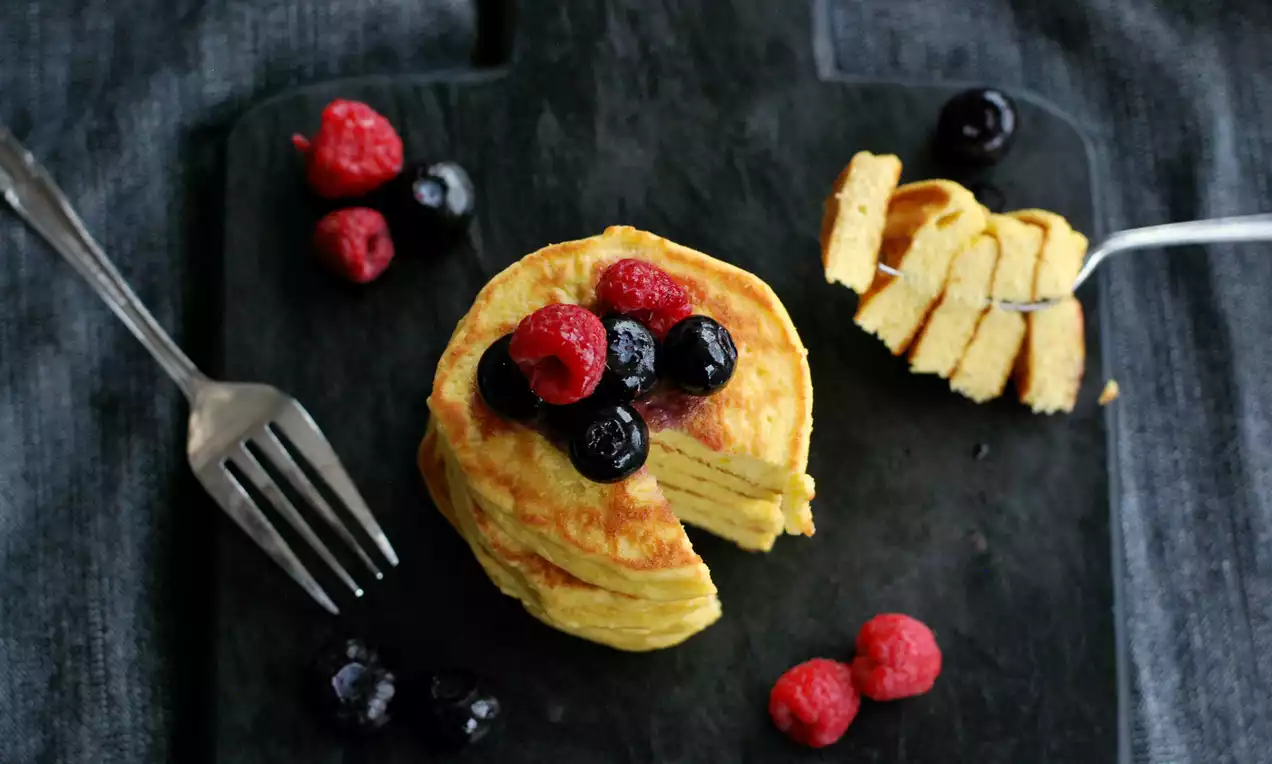 Hög saffransgul pannkaksstapel med blåbär och hallon på mörk tallrik, en bit uppträdd på gaffel och bär spridda runtom