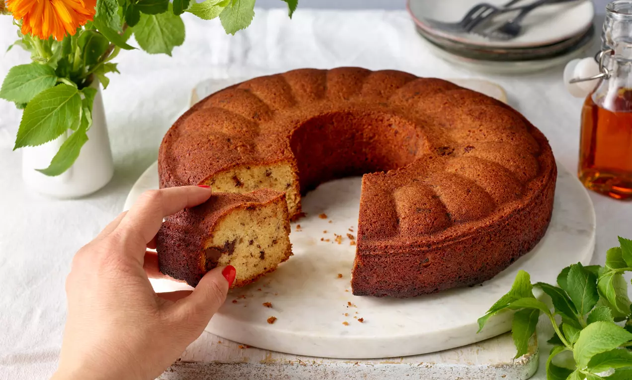 Ein goldbrauner Rumkranz mit strukturierter Oberfläche, angeschnitten auf weißer Platte, umgeben von frischem Grün und gelber Blüte