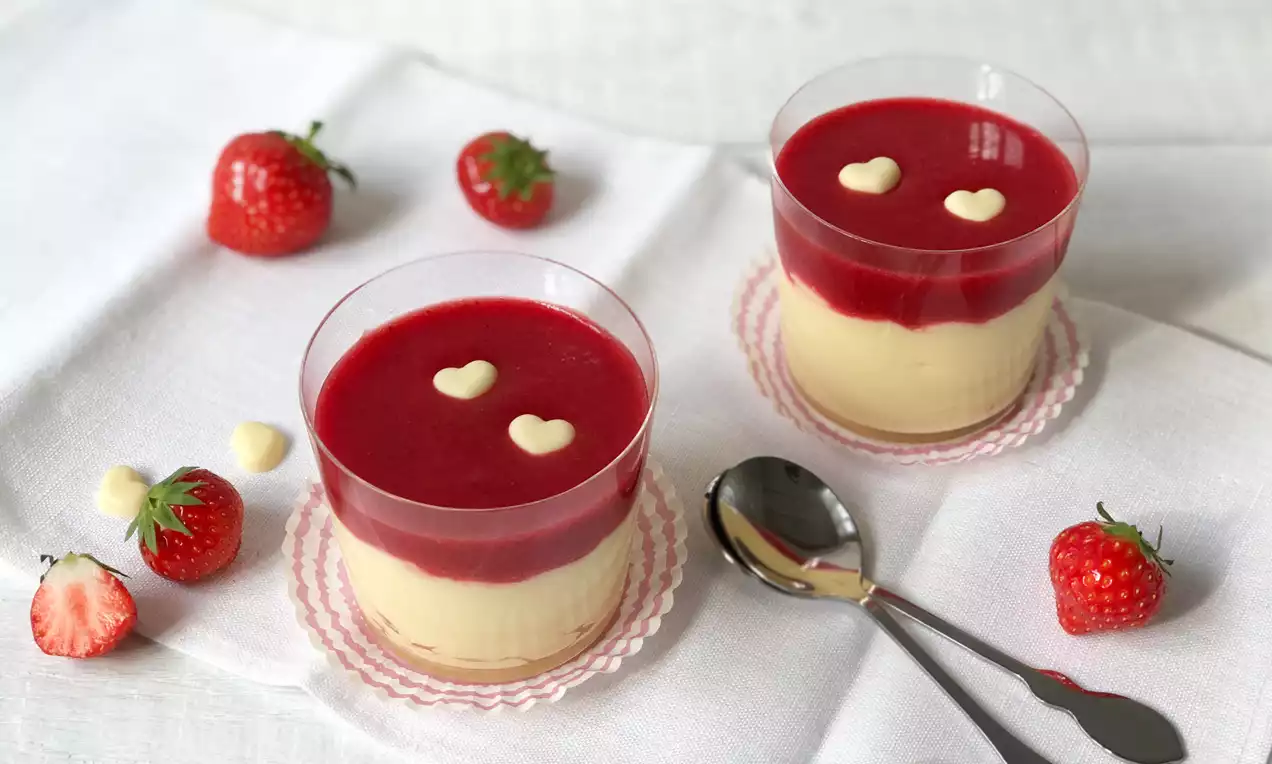 Feiner, hellgelber Pudding in klaren Gläsern, bedeckt mit roter Erdbeersoße und weißen Schokodekoren, arrangiert auf weißem Baumwolltuch