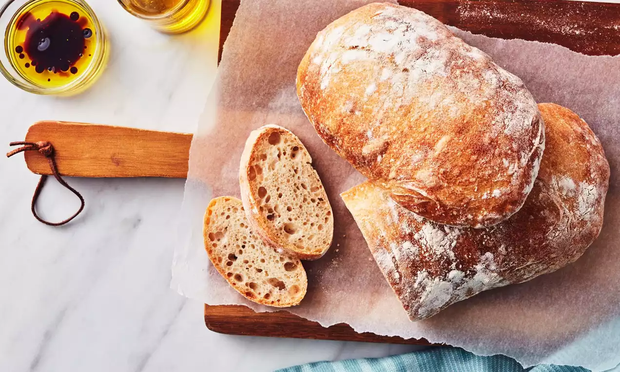 Two golden-brown ciabatta loaves with flour-dusted crusts on parchment paper, one sliced to reveal an airy crumb, on a wooden board