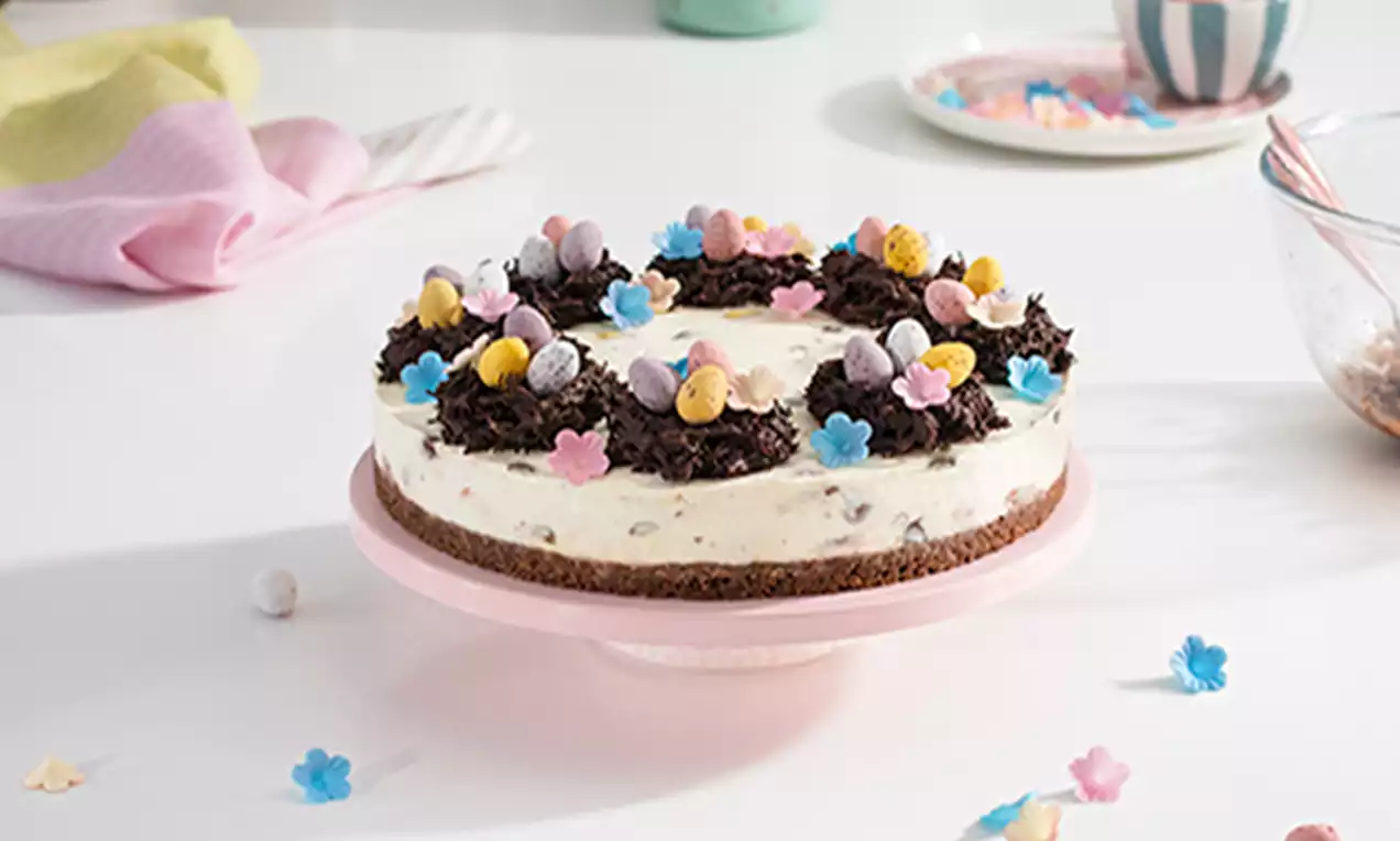 Creamy cheesecake with smooth filling on a brown biscuit base, topped with chocolate nests, pastel candy eggs, and pink and blue flower decorations