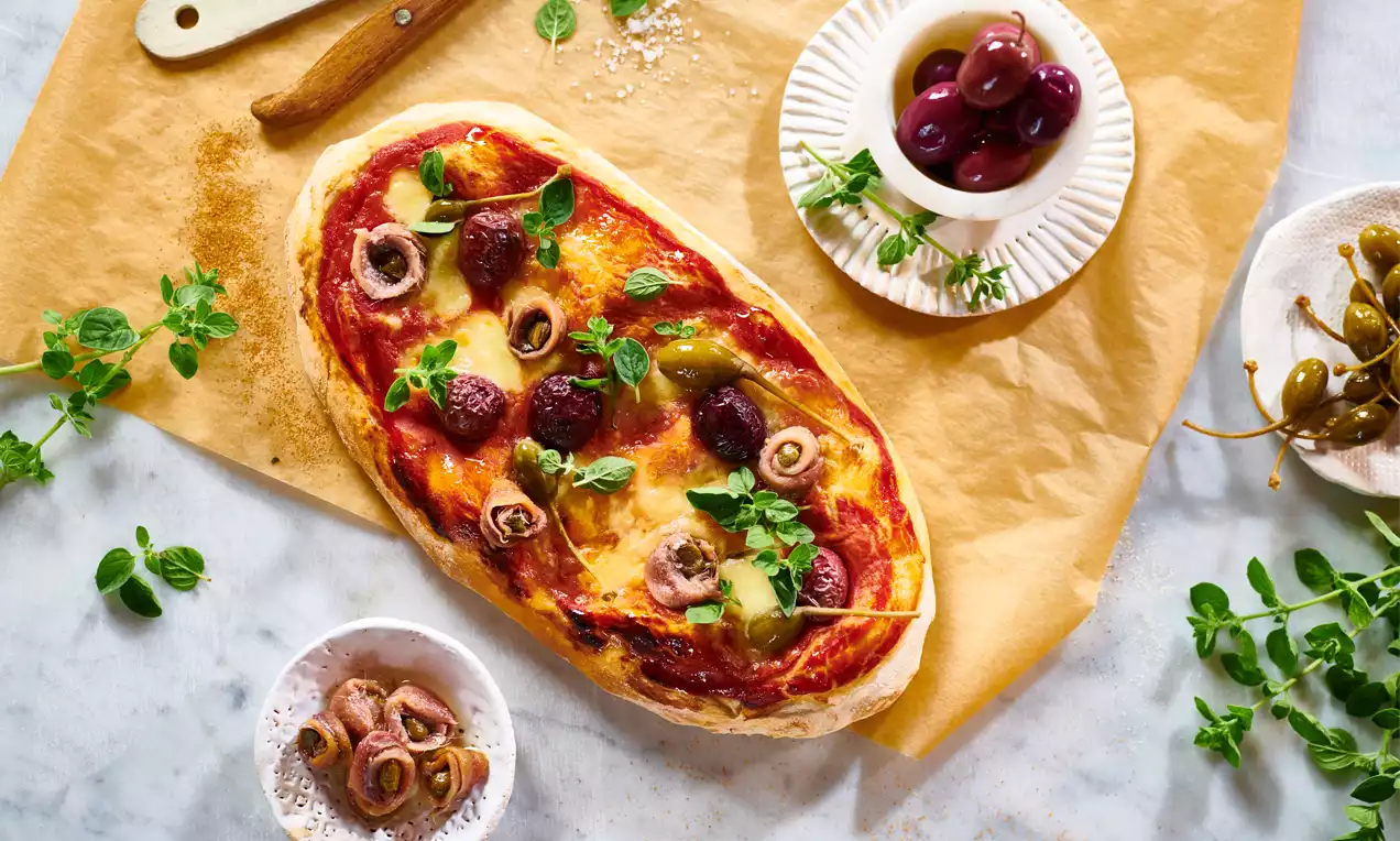 Pinsa mit ovaler Form, einzeln auf braunem Papier, mit leuchtend roter Tomatensauce, geschmolzenem Käse, Oliven, Sardellen und frischem Oregano
