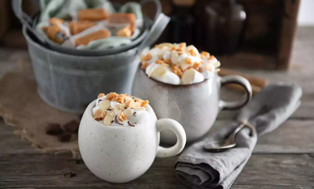 Krémová horúca čokoláda v keramických šálkach, snehobiela šľahačka posypaná zlatistými kúskami karameliek na drevenom stole