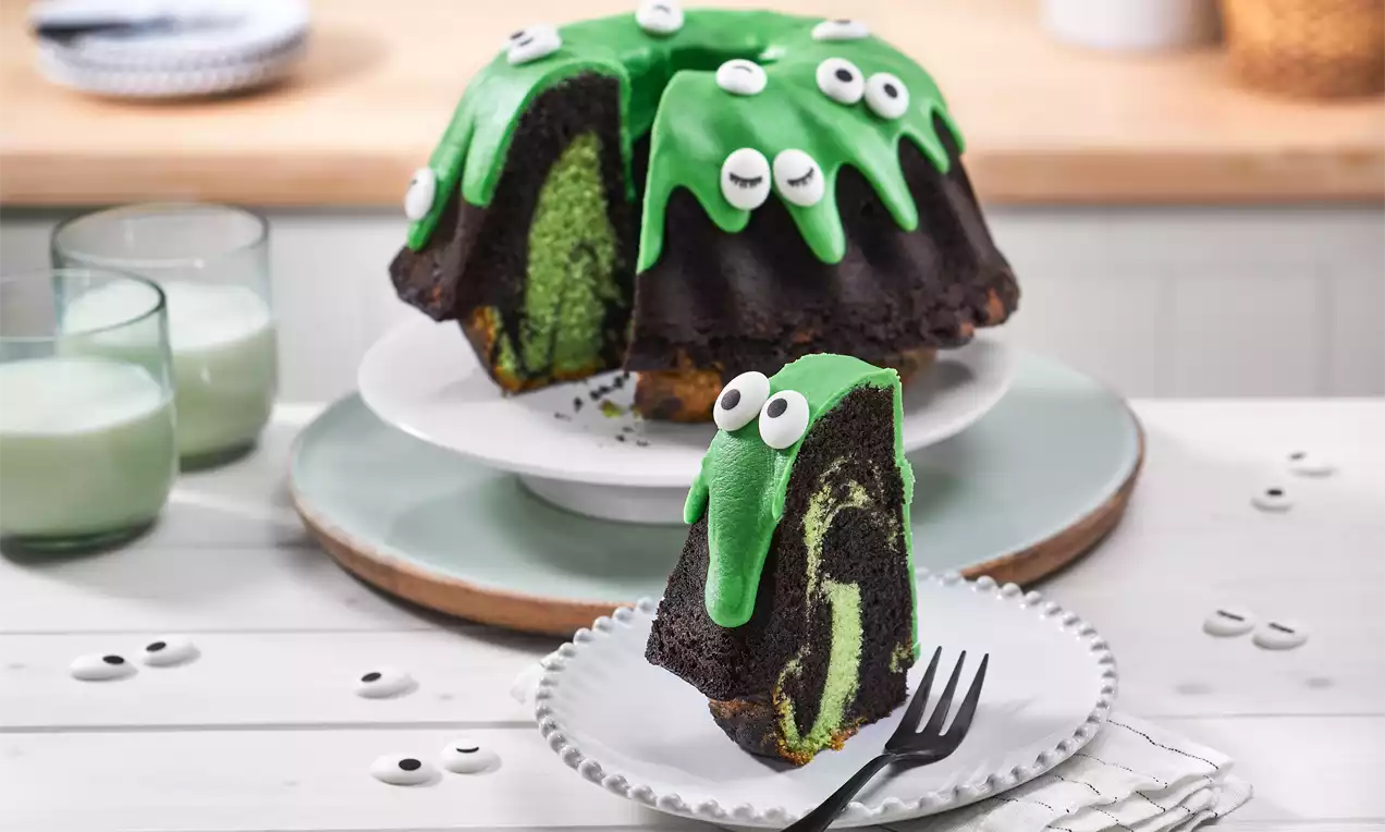 Schokoladiger Gugelhupf mit grünem Marmormuster, überzogen von giftgrünem Zuckerguss und dekoriert mit weißen Zuckeraugen auf einer Tortenplatte 