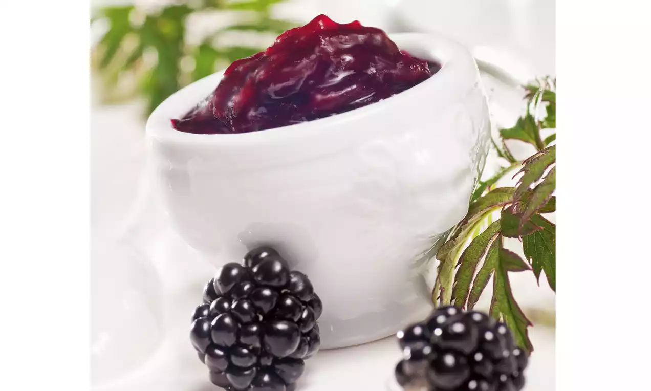 Donkerpaarse braambessenjam in een wit porseleinen kommetje, met glanzende zwarte braambessen en groene bladeren op de voorgrond