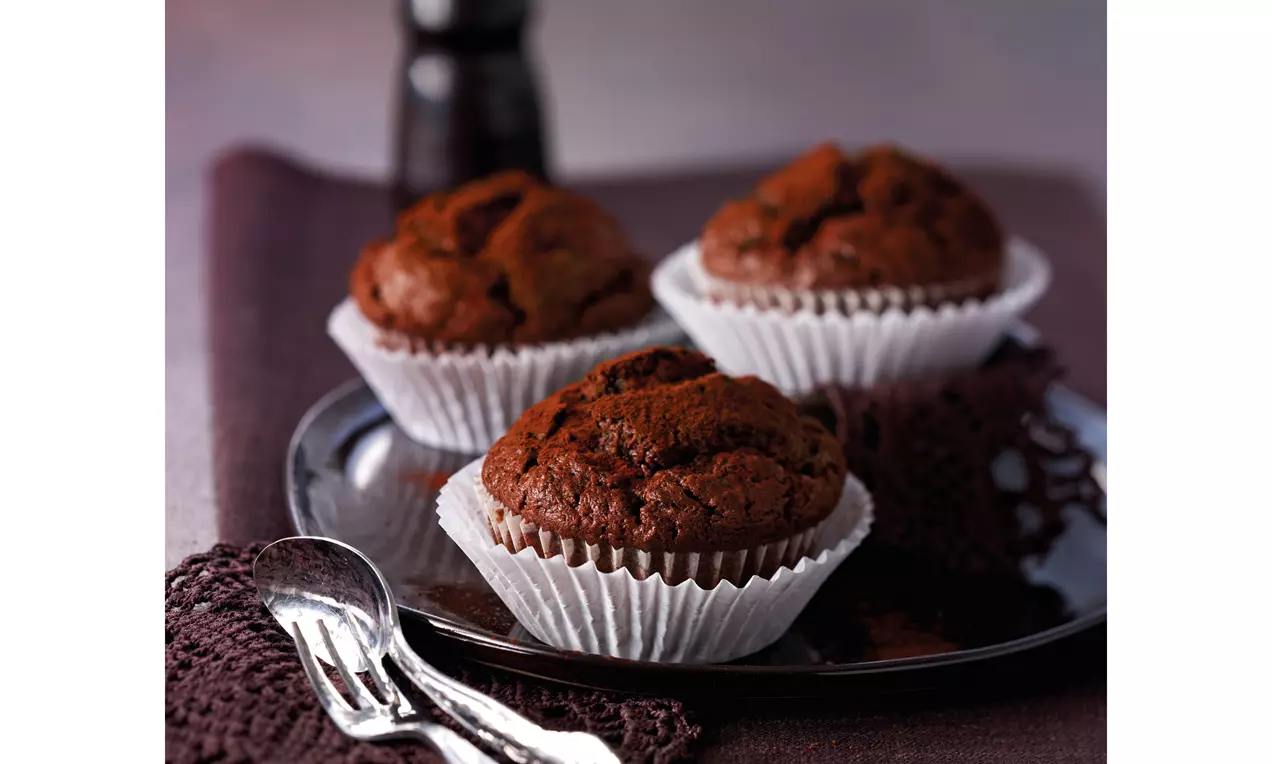 Dunkelbraune, lockere Schokoladen-Zucchini-Muffins in weißen Papierförmchen auf einem glänzenden Teller mit lilafarbenem Stoff drapiert