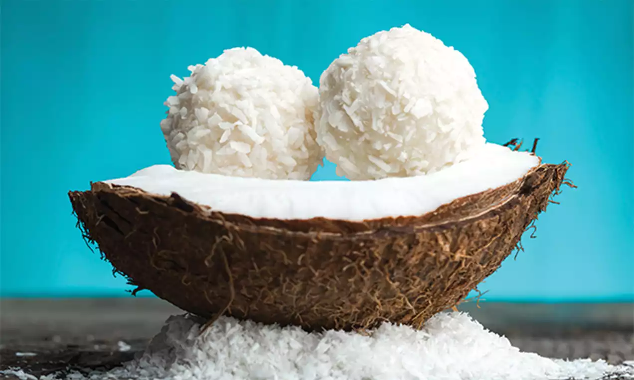 Two white coconut cake pops with delicate flakes sit atop creamy coconut flesh inside a rustic brown coconut shell on a bed of grated coconut