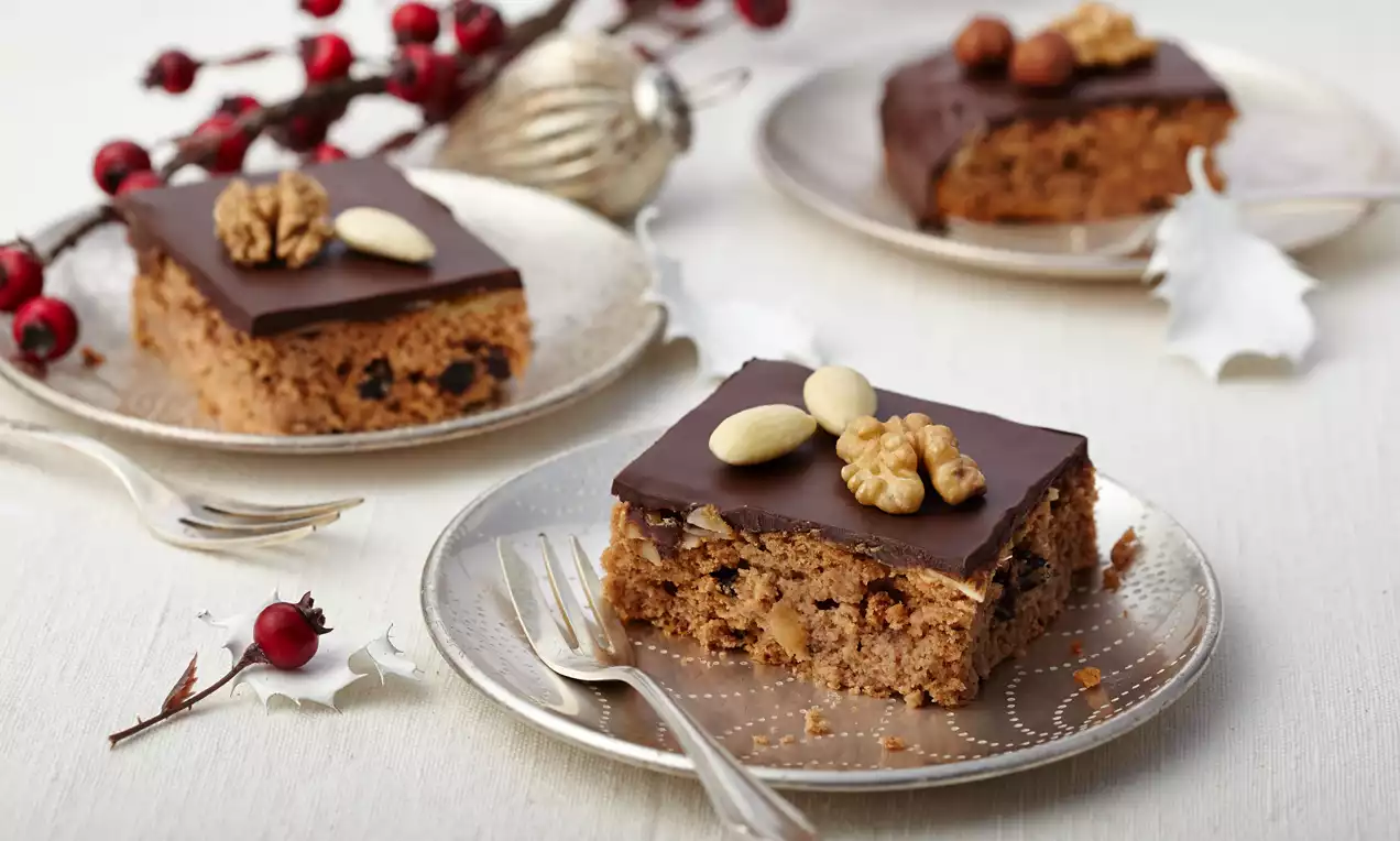 Honigkuchen mit dunkler Schokoladenglasur, Walnuss und Mandeln auf silbernem Teller, arrangiert mit roten Beerenzweigen und Deko