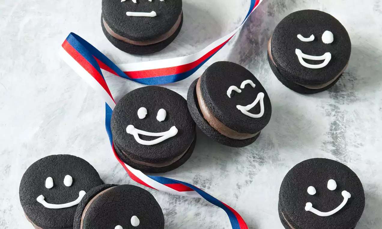 Černá kulatá linecká kolečka s čokoládovou náplní, zdobená bílými veselými tvářičkami, na šedém povrchu s trikolórovou stuhou