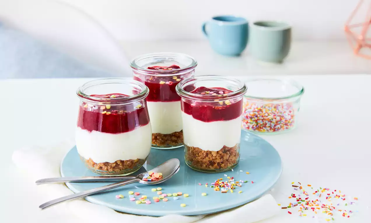 Cheesecake-Dessert im Glas mit braunem Keksboden, weißer Creme, rotem Fruchtspiegel und bunten Zuckerstreuseln auf einem hellblauen Teller