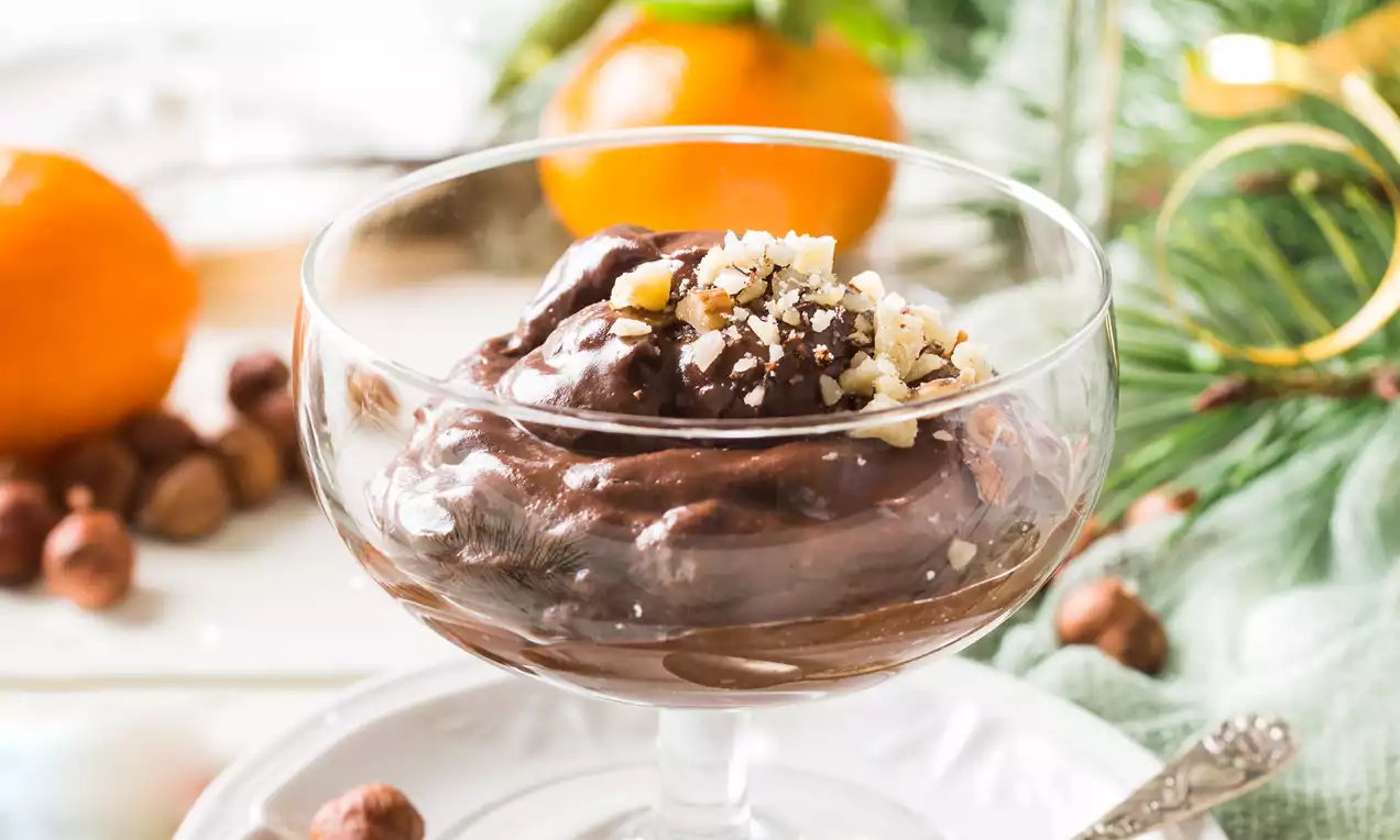 Romige chocolademousse in een glas, gegarneerd met gehakte noten, geplaatst op een wit bord met mandarijnen en dennentakken op de achtergrond