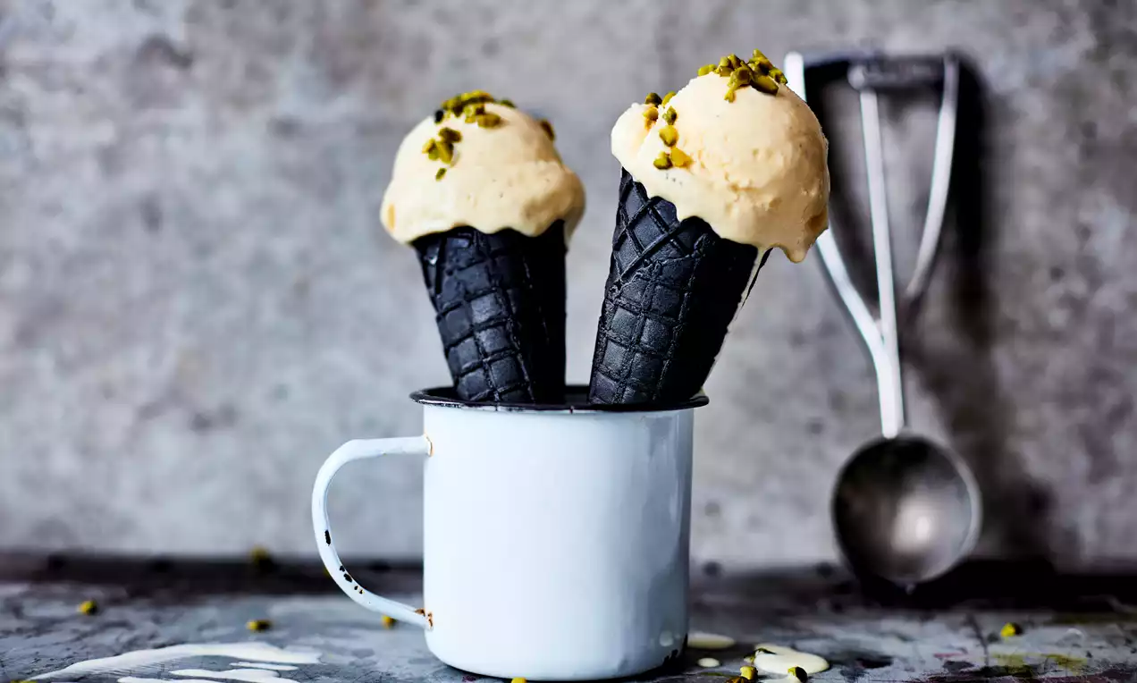 Zelfgemaakt vanille-ijs met romig beige bolletjes op zwarte hoorntjes in een lichtblauw metalen mok, gegarneerd met groene pistachenootjes