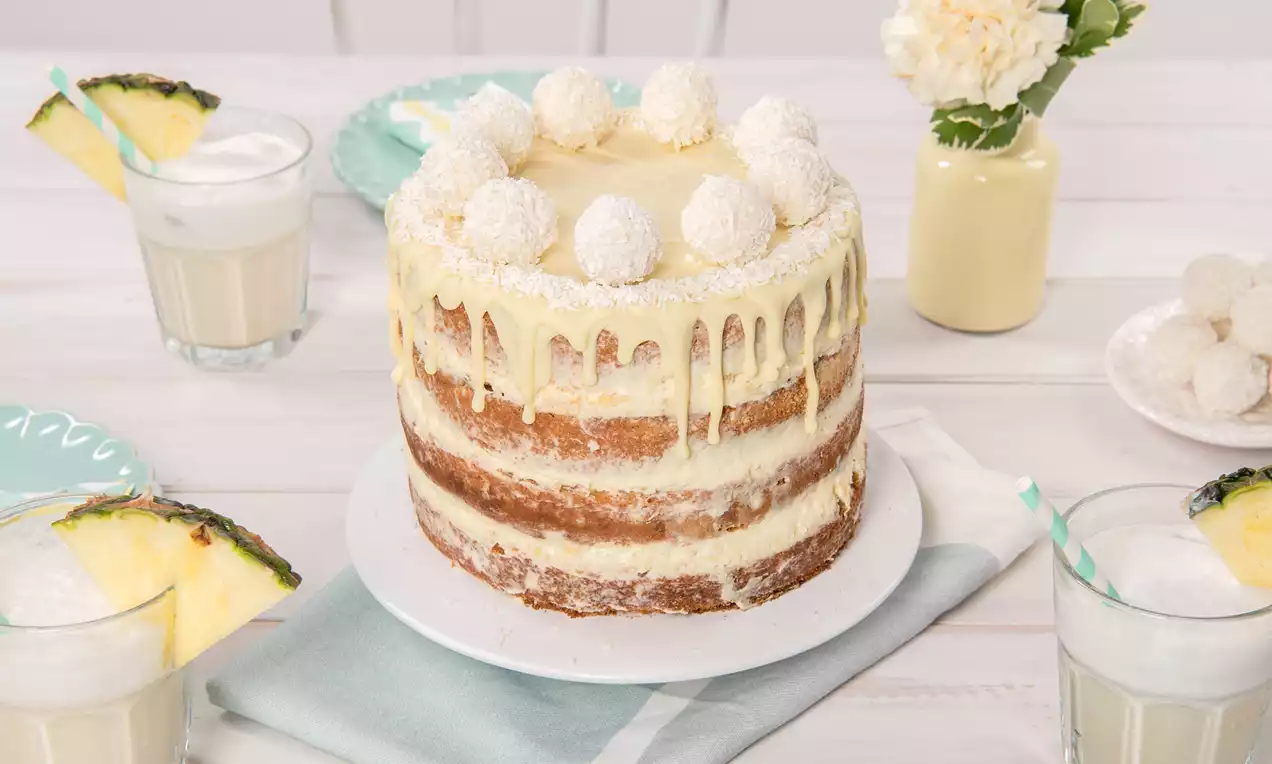 Pina-Colada-Torte mit goldbraunem Schichtboden, heller Creme, weißer Glasur und Kokoskugeln auf weißem Teller mit tropischen Getränken