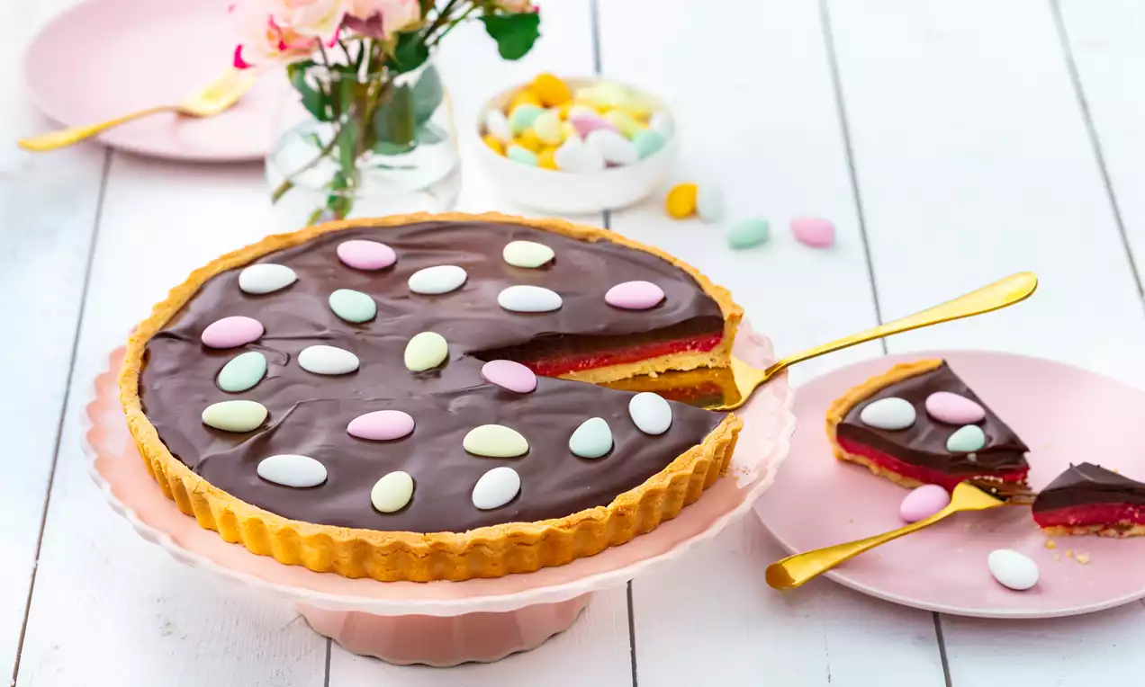 Runde Schokoladen-Himbeer-Tarte mit glänzender dunkler Glasur und bunten Dragees auf pastellfarbenem Tortenständer, umgeben von rosa Geschirr