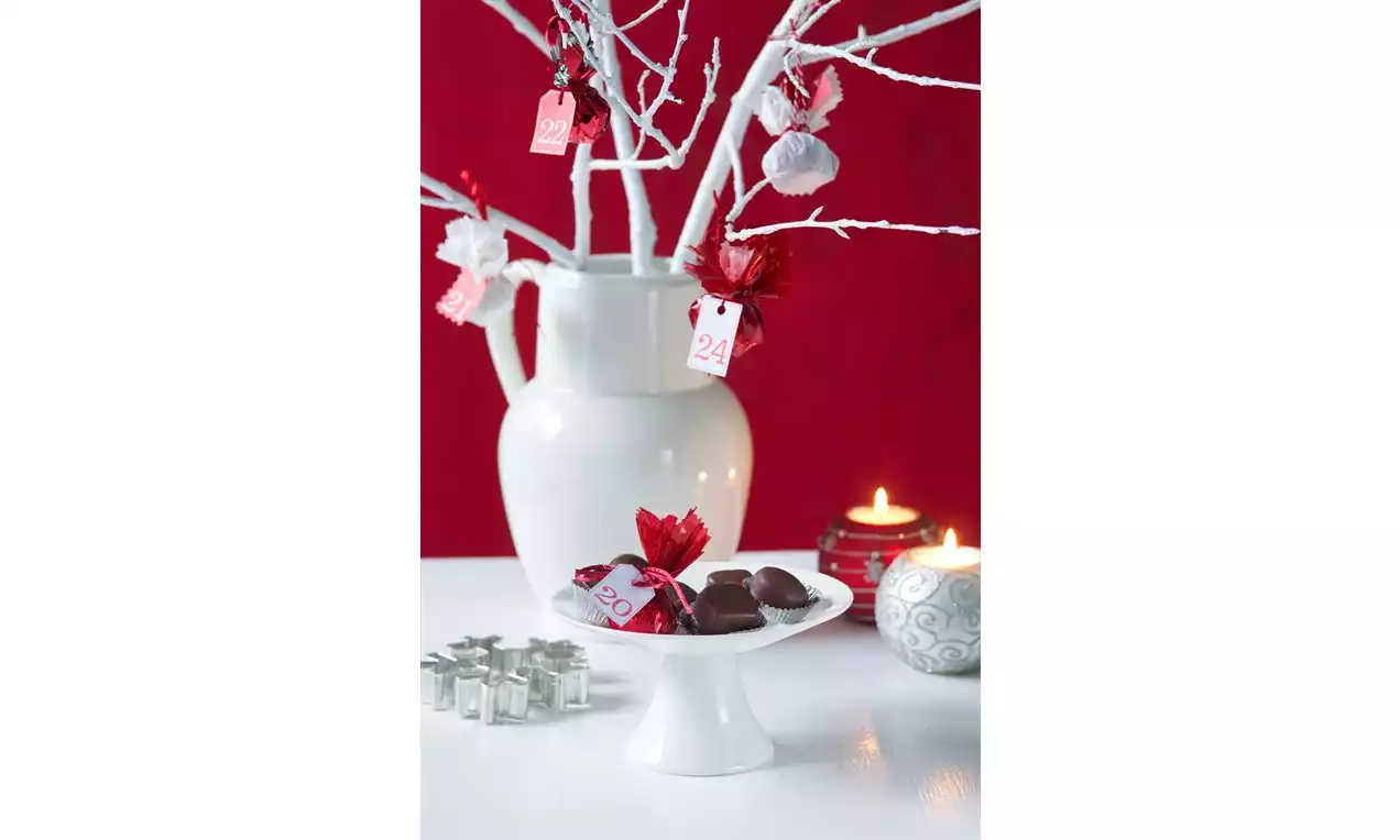 Čokoládové pralinky na bielom stojane obklopené červenými ozdobami, vedľa sviečok a stromčekových dekorácií v kontraste s červeným pozadím