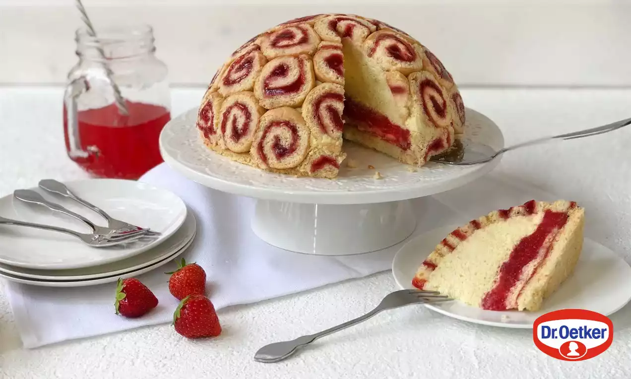 Světle béžový dort ve tvaru polokoule tvořený spirálovými plátky rolády s červenou jahodovou náplní na bílé podložce s čerstvými jahodami