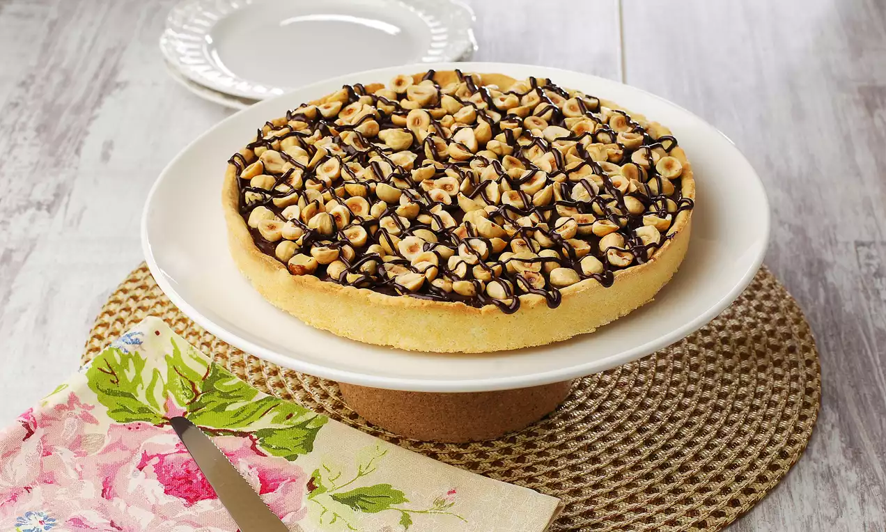 Altın rengi tart tabanı üzerinde bütün fındıklarla kaplanmış ve ince çikolata şeritleriyle süslenmiş, beyaz bir ayaklı tabakta sunulmuş.