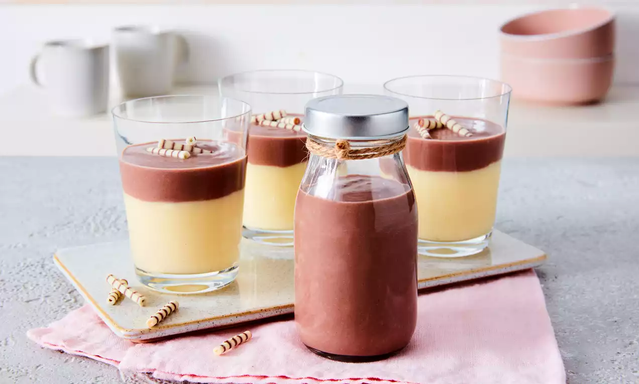 Vier Gläser mit gelber Vanillecreme und dunkelbraunem Schokoladenpudding, garniert mit Schokoröllchen, neben einer Flasche mit Schokosoße.