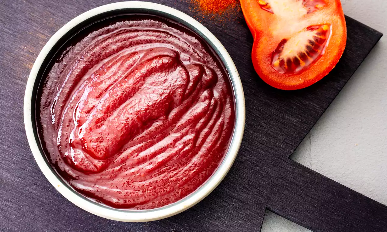Hot BBQ sauce in a white bowl with glossy deep red texture, set on a dark board with sliced tomato, peppercorns and paprika powder