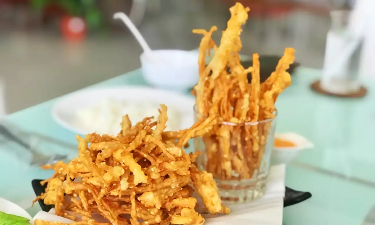 Golden crispy enoki mushrooms with thin, delicate strands, piled on white parchment and served in a clear glass against a light table setting