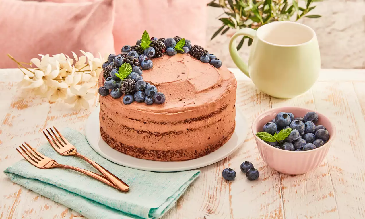 Halbhoher Semi-Naked-Cake mit brauner Creme, garniert mit Blaubeeren, Brombeeren und Minzblättern auf weißem Teller, dekoriert mit Blumen