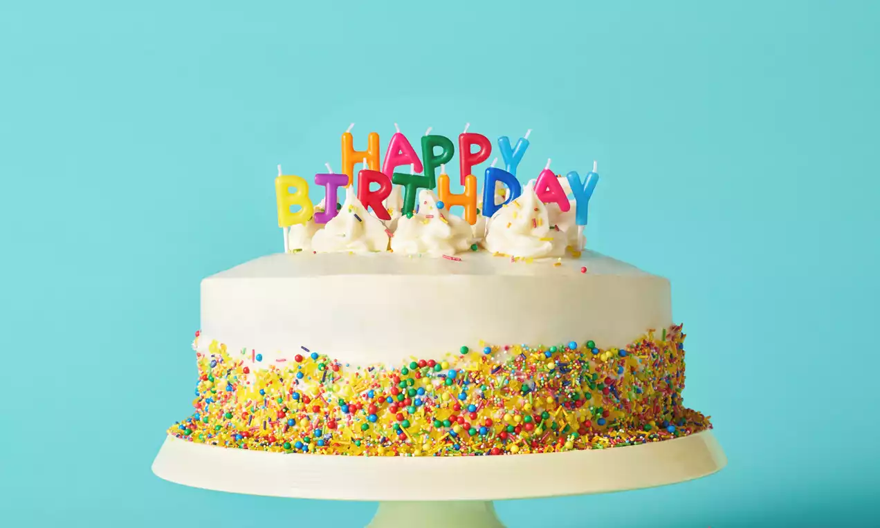 Vanilla buttercream birthday cake with smooth white icing, colourful sprinkles coating the sides, topped with candles spelling Happy Birthday on a cream pedestal