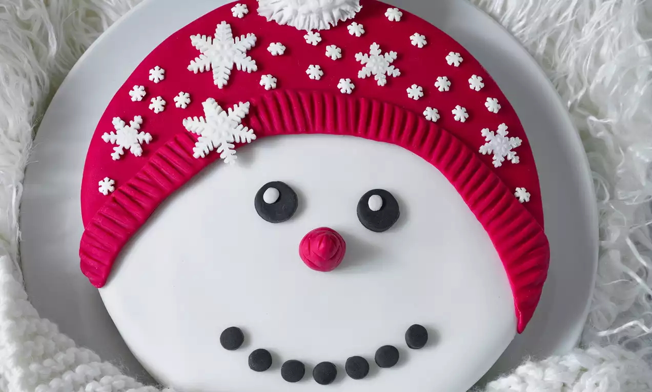 Biela torta v tvare snehuliaka so zvýraznenou červenou čiapkou, ozdobená malými snehovými vločkami, čiernymi očami a úsmevom z bodiek