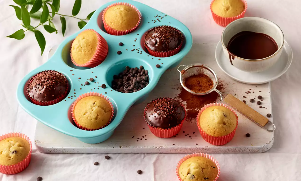 Goldene und schokoladenbraune Muffins mit weicher Oberfläche, teils mit Schokostücken, in roten Förmchen auf einem Tablett mit Kakao und Kaffee