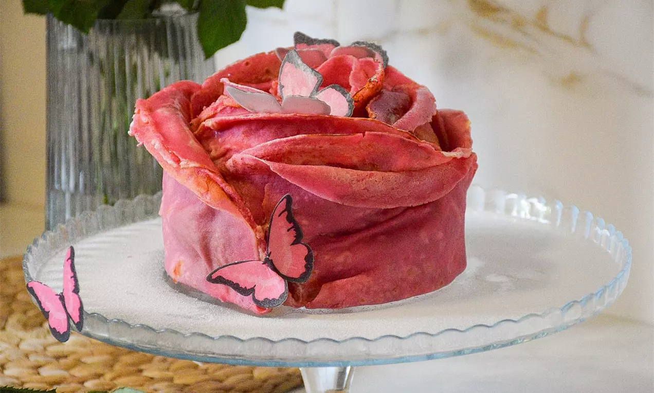 Torta ružičastih nijansi s teksturom nalik laticama, ukrašena papirnim leptirima na staklenom tanjuru, s pozadinom zelenog lišća