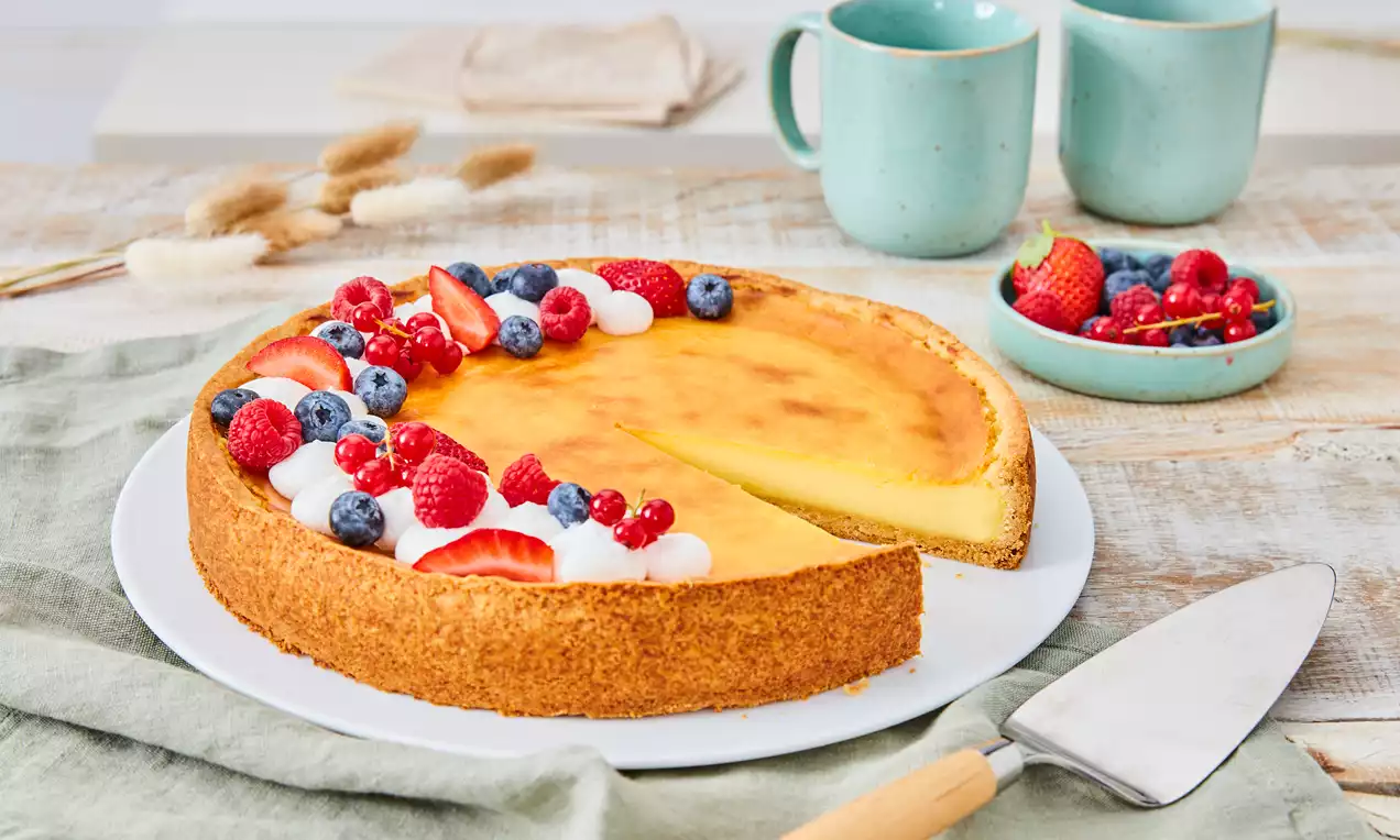 Veganer Käsekuchen mit goldgelber Oberfläche, auf weißem Teller präsentiert, garniert mit Sahnetupfen, roten und blauen Beeren