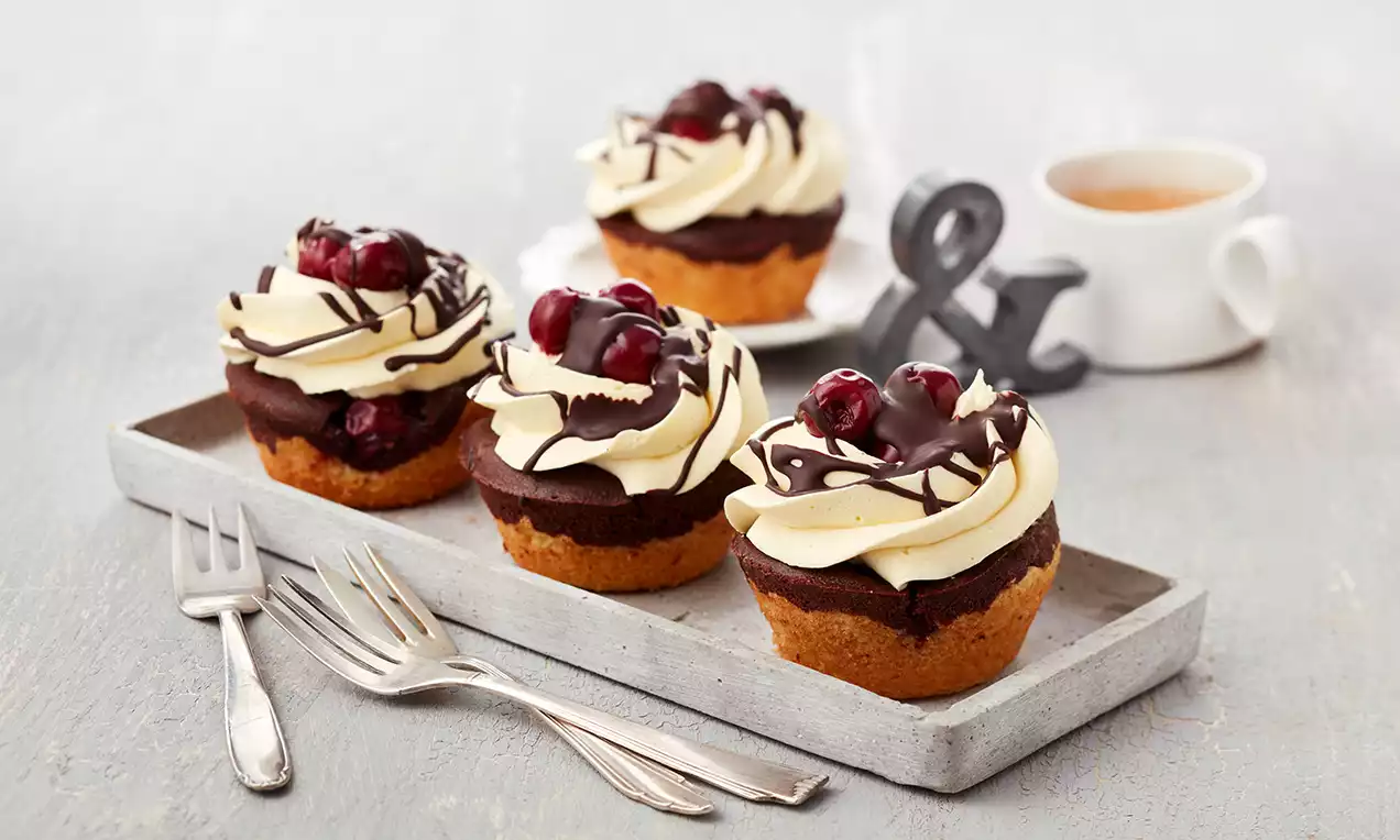 Donauwellen-Muffins mit hellgelber Creme und Schokoglasur, garniert mit roten Kirschen, auf grauem Tablett mit Gabeln angerichtet