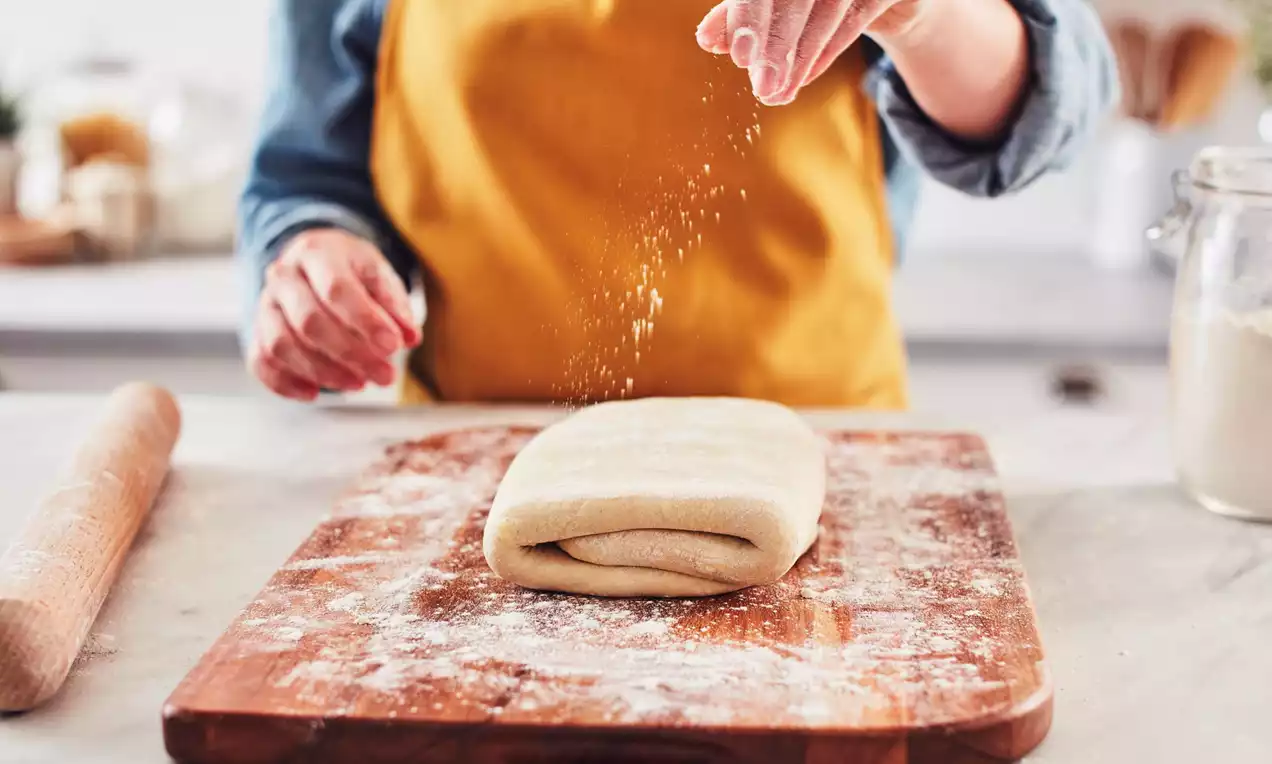Folded puff pastry dough with smooth, pale surface on a floured wooden board, sprinkled with flour by a hand in mid-motion