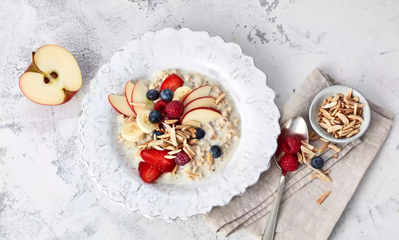 Cremiger Porridge mit Apfelscheiben, Bananenscheiben, Himbeeren, Blaubeeren und gerösteten Mandeln auf einem weißen Teller angerichtet