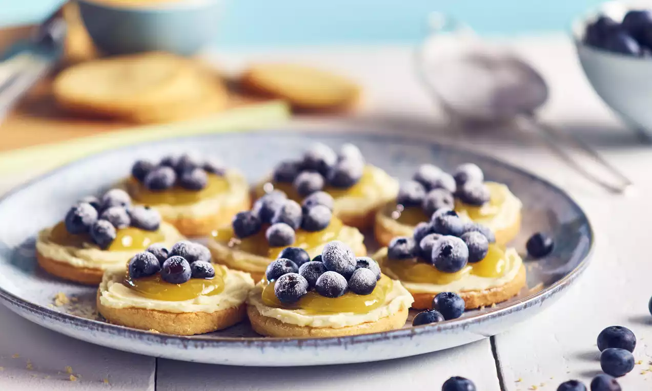 Round cookies with a golden base topped with pale yellow lemon cream, glossy blueberries, and a dusting of icing sugar on a blue plate