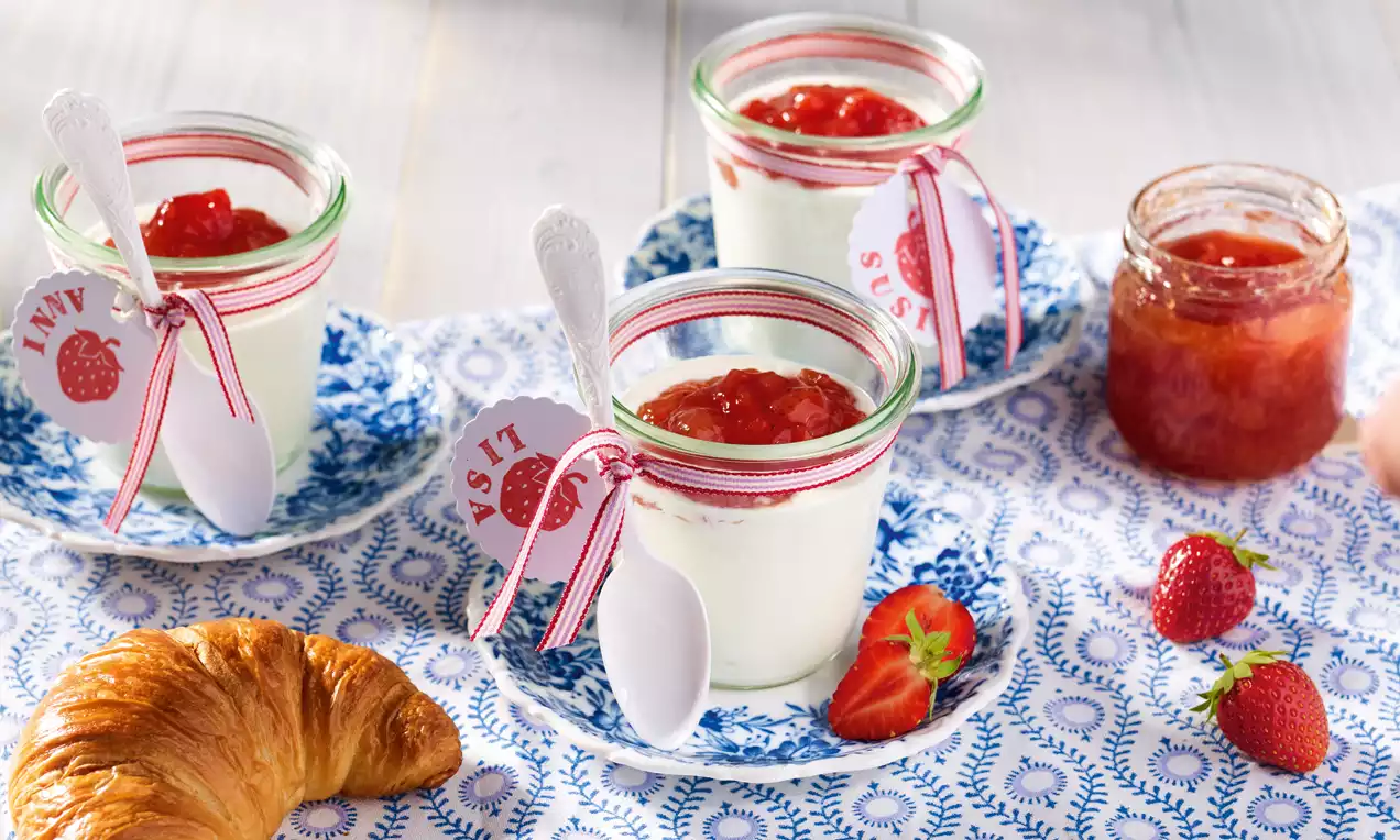 Lágy fehér joghurt üvegekben piros eper-grépfrút lekvárral, mintás tányérokon díszkötéssel, friss eper és aranybarna croissant mellett