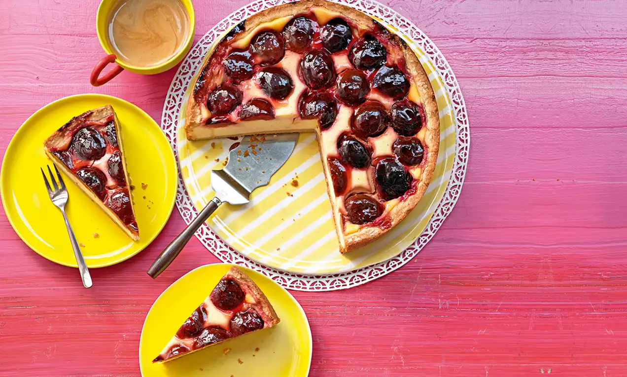 Zwetschgen-Schmand-Kuchen mit glänzend dunkelroten Zwetschgenhälften auf heller Creme, serviert auf gelbem Teller und rosa Holzuntergrund