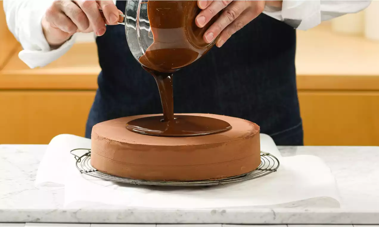 Dunkle Schokolade aus einem Glasgefäß fließt gleichmäßig auf eine runde, glatte Torte mit brauner Creme auf einem Gitter über weißem Papier