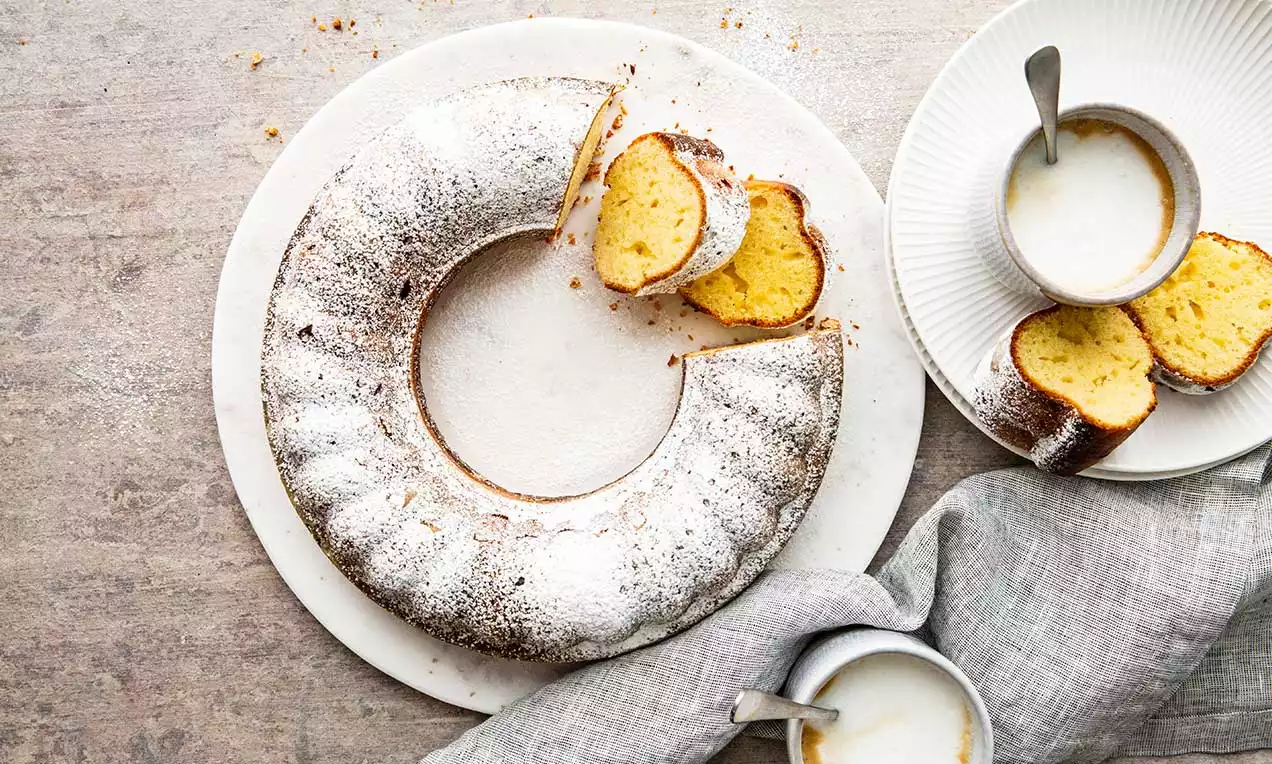 Ciambella dorata e soffice cosparsa di zucchero a velo bianco, servita su un piatto con morbide fette e tazze di caffè affiancate