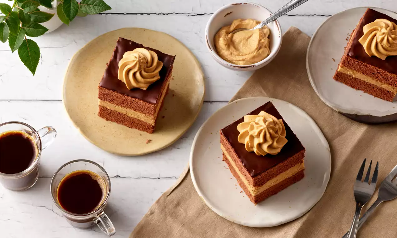 Mokkawürfel mit dunkler Glasur, cremigem Kaffeetopping und geschichteten Teiglagen auf einem weißen Teller neben Kaffee und Dekoration
