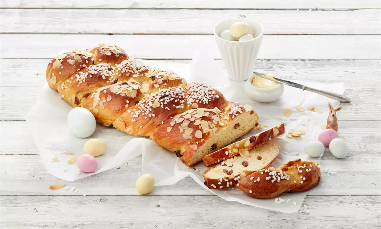 Osterzopf sa zlatnosmeđom površinom, posut listićima badema i šećerom, delikatno narezan, uz ukrasne obojene jaja na bijeloj podlozi