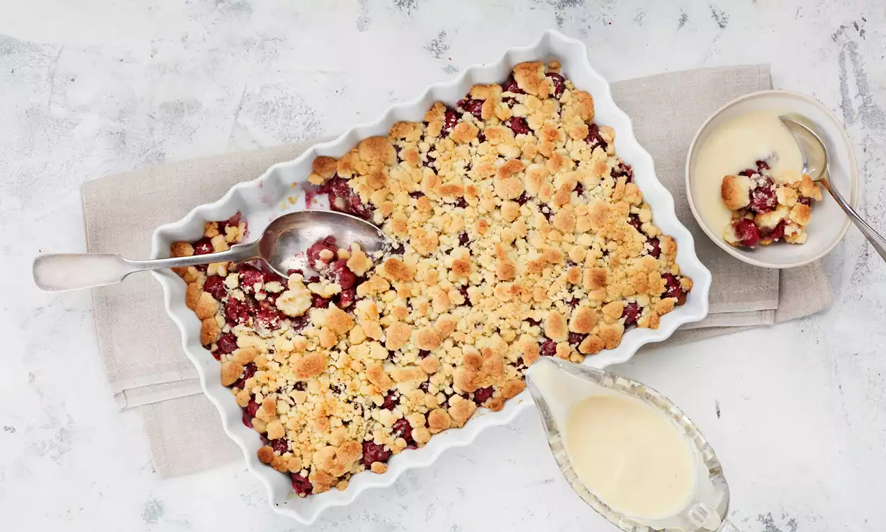 Goldbrauner Kirsch-Crumble mit fruchtiger Füllung in rechteckiger Form, daneben eine helle Sauce in Kännchen und eine Portion in Schale